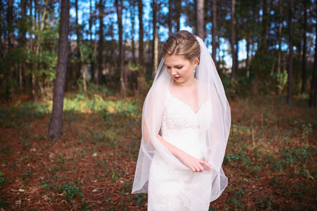 young woman on her wedding day wearing a veil and a white wedding dress looking down at the ground with trees in the background