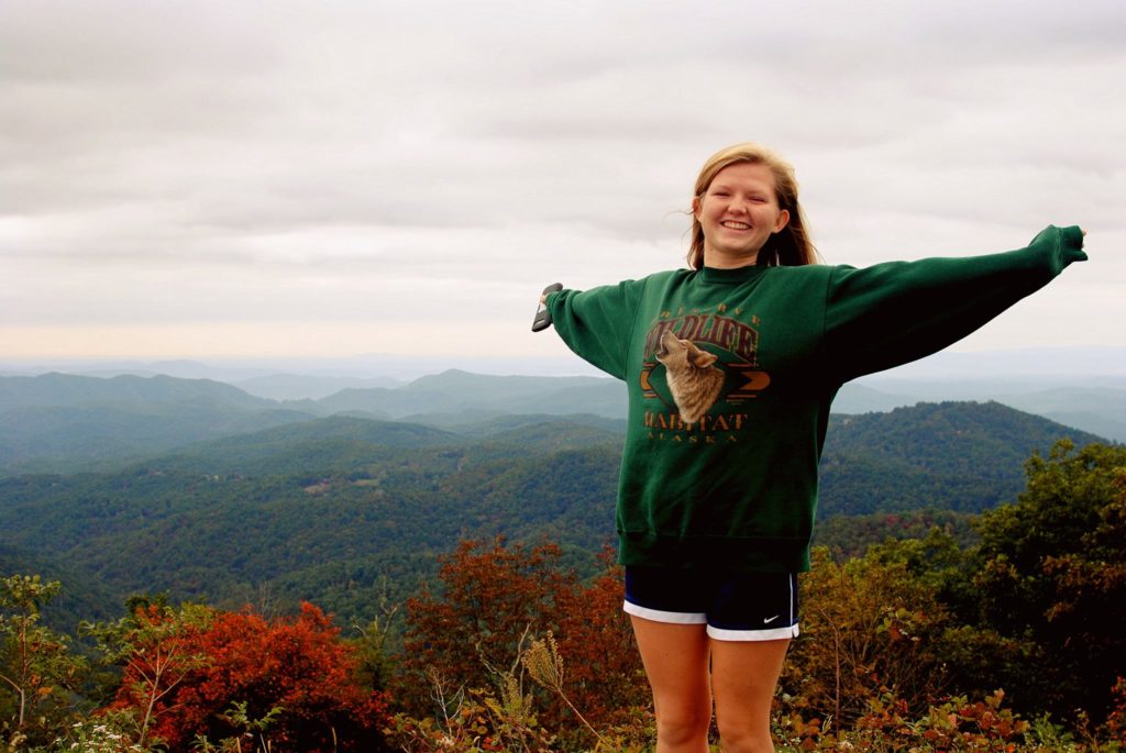 young woman with arms spread out with the mountains in the background wearing a sweatshirt and shorts smiling