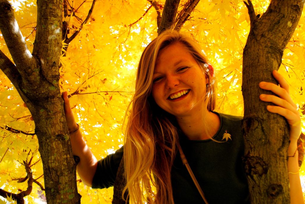 young healthy looking girl smiling in yellow tree with bright yellow fall colors, the girl has a green sweater on and long blonde hair